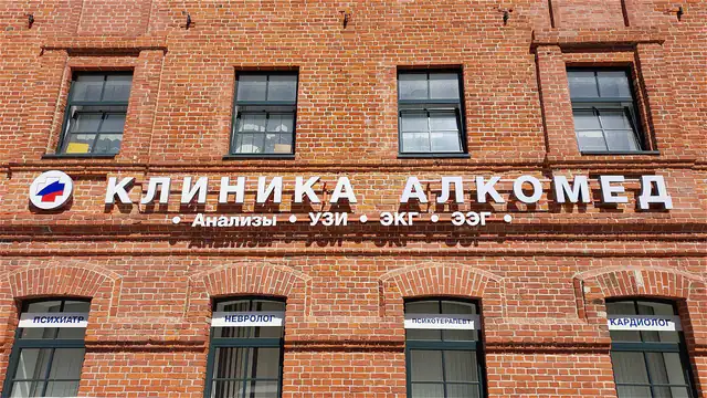 Здание клиники снаружи: краснокирпичный фасад с вывеской «Клиника АлкоМед», у окон таблички — психиатр, невролог, психотерапевт, кардиолог